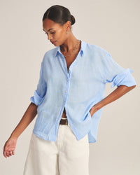 A woman with dark hair in a low bun wears the Velvet by Graham & Spencer Brighton Long Sleeve Button Up in Sailboat with rolled sleeves and white pants. She stands against a neutral background, looking down with one hand on her hip.