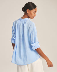 A woman with dark hair in a low bun wears the Velvet by Graham & Spencer Brighton Long Sleeve Button Up in Sailboat and white pants, turned slightly away and looking over her shoulder against a plain background.