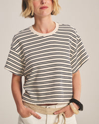 A person with short blonde hair wears the Brissa Short Sleeve Crew Neck Top in Natural/Navy by Velvet by Graham & Spencer, light pants with a beige rope belt, and chunky bracelets, standing hands in pockets against a plain background.