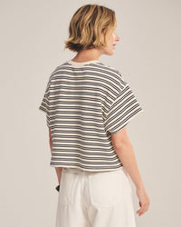 A person with short blond hair is wearing the Brissa Short Sleeve Crew Neck Top in Natural/Navy by Velvet by Graham & Spencer and white pants, standing and facing away from the camera against a plain background.