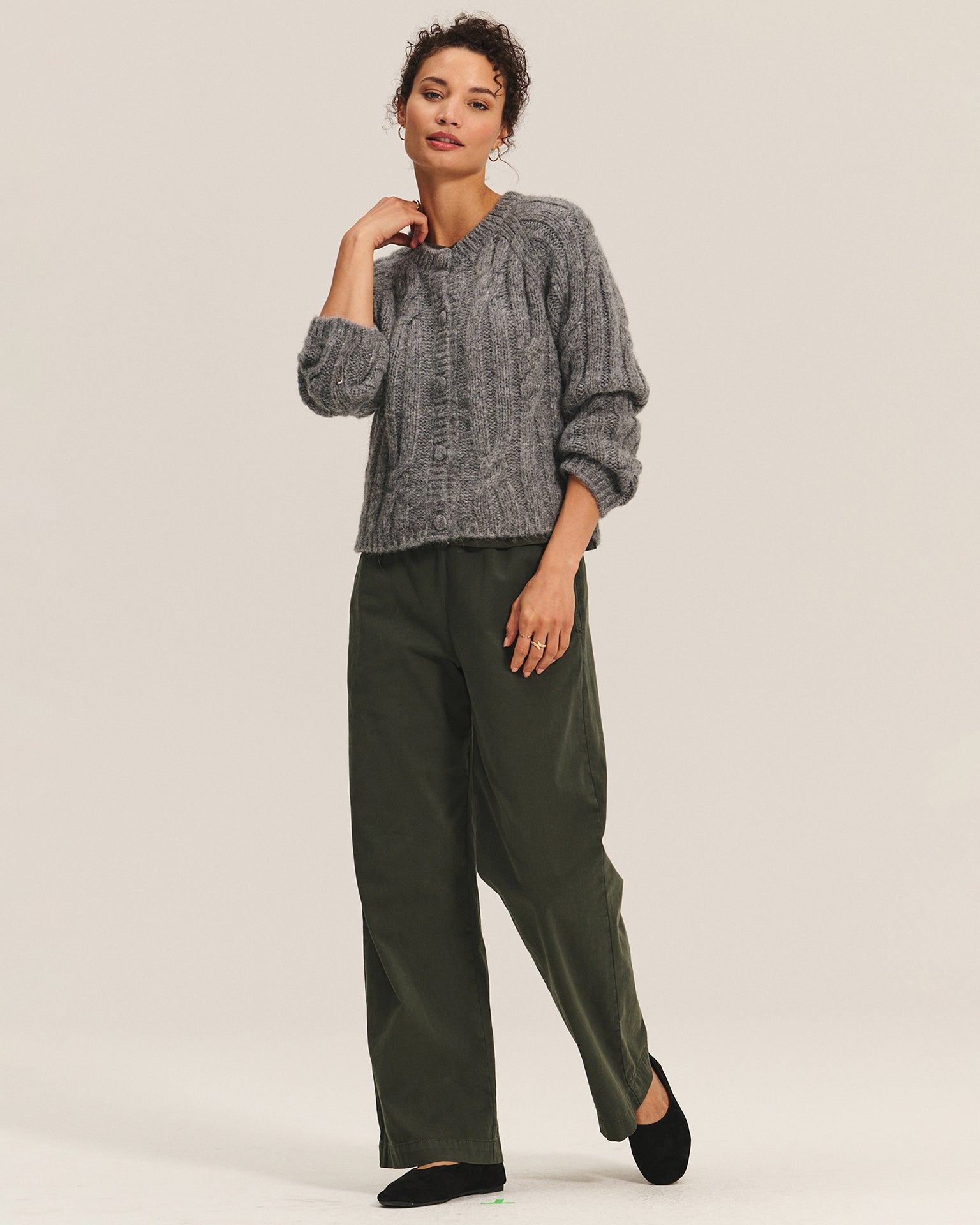 A woman stands against a plain background wearing the Velvet by Graham & Spencer Allison Long Sleeve Crew Cardi in Heather Grey, paired with loose olive green pants and black shoes. She has curly hair tied back, posing with one hand on her neck and the other in her pocket.