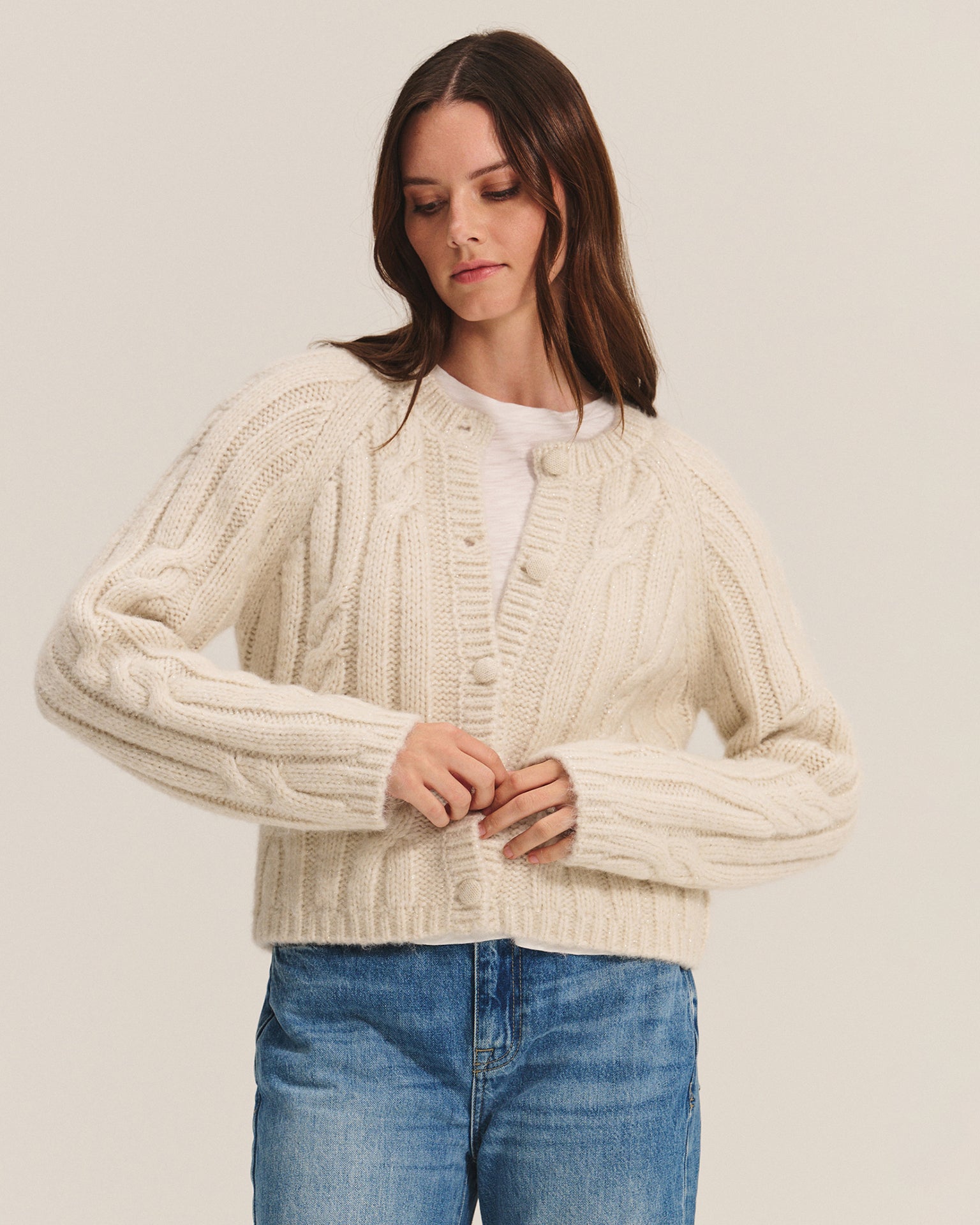 A woman with long brown hair wears the Allison Long Sleeve Crew Cardi in Ivory by Velvet by Graham & Spencer over a white t-shirt and blue jeans, standing against a plain light background.