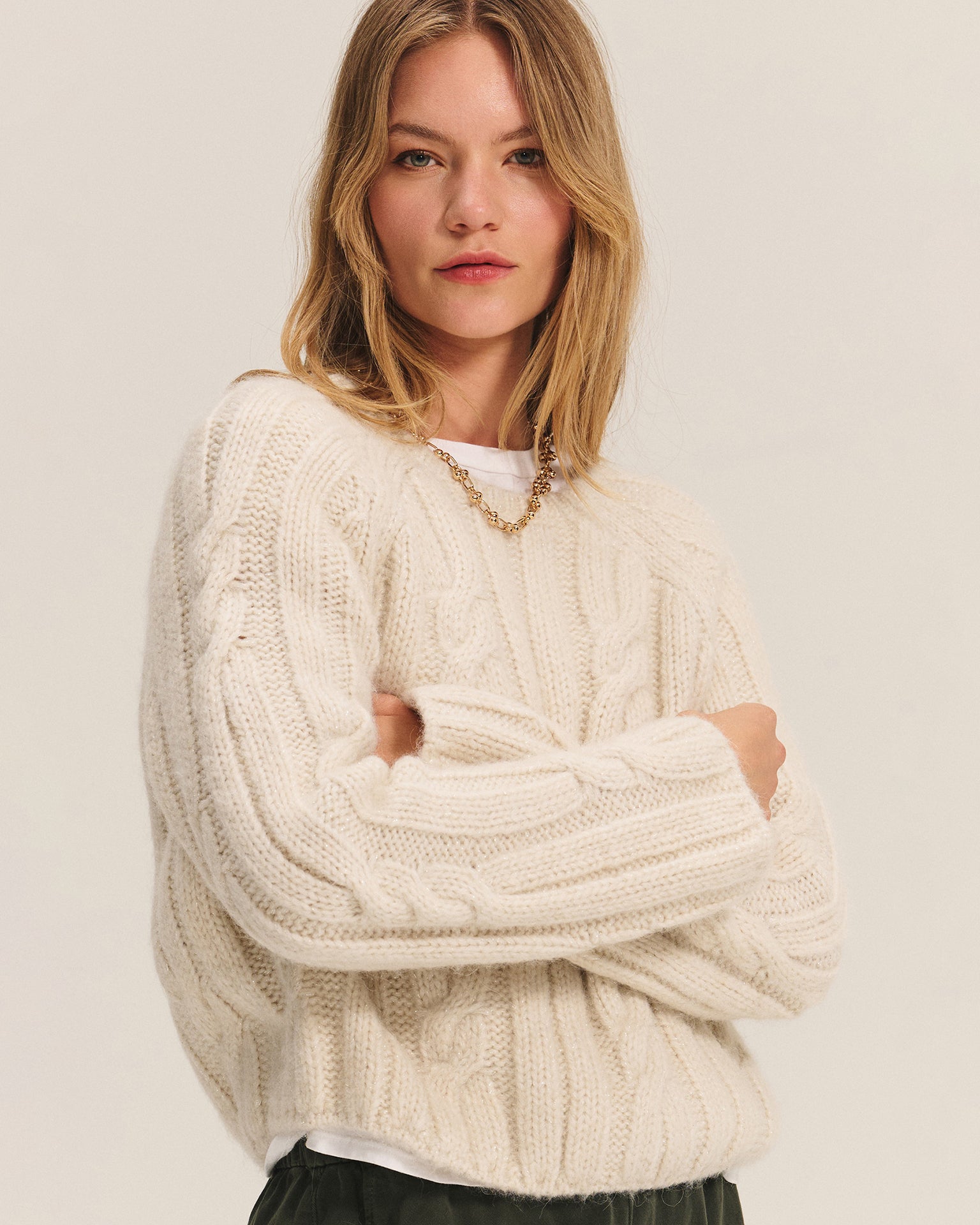 A woman with long blonde hair wears the Velvet by Graham & Spencer Marina Long Sleeve Crew Top in Ivory over a white shirt and gold chain necklace. She stands with arms crossed, looking confidently at the camera against a plain background.