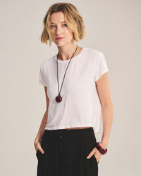 A woman with short blonde hair wears the Casey Short Sleeve Crew Neck Top in White by Velvet by Graham & Spencer, paired with black pants, a large round pendant necklace, and a chunky bracelet. She stands hands in pockets against a plain background.