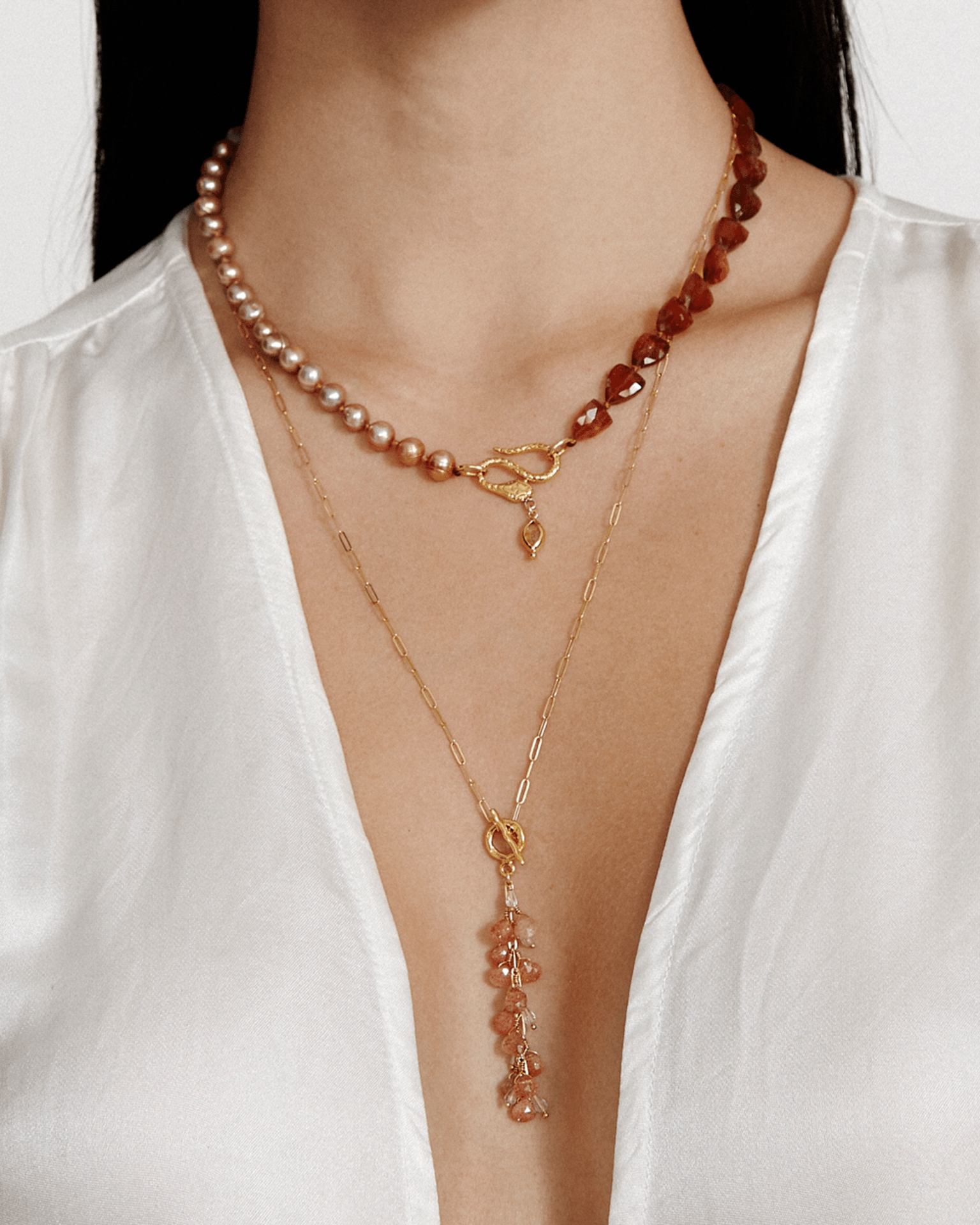 A person wears a Pacifica Necklace in Sunstone Mix by Chan Luu with layers featuring pearls, red beads, and an 18k gold plated sterling silver necklace with a dangling aquamarine multi-gem pendant, perfectly paired with a white V-neck top.