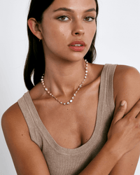A woman with long brown hair wears a sleeveless beige ribbed top and the Chan Luu Daphne Beaded Necklace in White Mix. She looks at the camera with a neutral expression, one arm crossed over her chest, against a plain light background.