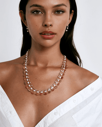 A woman in a white off-the-shoulder shirt wears layered pearl and pink bead necklaces, including the Chan Luu Santa Fe Necklace in Sunstone, paired with matching pearl drop earrings, posing against a plain background.