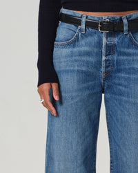 A person wears the Citizens of Humanity Annina High Rise Wide Leg 30in in Sunland jeans, styled with a black belt and dark long-sleeve top, showing rings on their left hand near their hip; image is cropped at waist and mid-thigh.