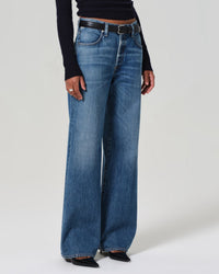 A person stands against a plain light background wearing Citizens of Humanity’s Annina High Rise Wide Leg 30in jeans in Sunland, paired with a black belt, long-sleeve black top, and black pointed-toe shoes. The upper body is partially out of frame.