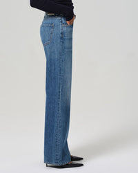A person wearing Citizens of Humanity Annina High Rise Wide Leg 30in in Sunland jeans and black pointed-toe shoes stands sideways with one hand in their pocket against a plain light background.