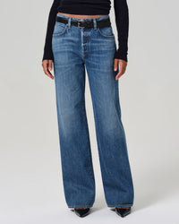 Wearing the Annina High Rise Wide Leg 30in in Sunland by Citizens of Humanity, paired with a black belt and black long-sleeve top, a person stands against a plain light background. Only their lower torso and legs are visible.