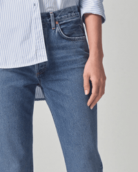 A person wearing Citizens of Humanity Charlotte High Rise Straight in First Class jeans stands against a plain, light background, with only their lower torso and hand visible.