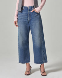 A person in Citizens of Humanity Gaucho Vintage Wide Leg jeans in Oasis, a light pink long-sleeve top, gray vest, and strappy heeled sandals stands with one hand in a pocket against a plain light background.