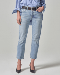 Wearing Citizens of Humanity’s Isla Low Rise Straight in Spector jeans, paired with a blue button-up shirt, black belt, and pointed-toe heels, a person stands against a plain light gray background.
