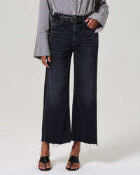 A person in a gray striped blouse, black belt, black open-toe sandals, and Citizens of Humanity Lyra Wide Leg Crop in Medallion jeans stands against a plain light background.
