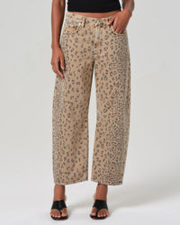 A person wears Citizens of Humanity’s Miro Relaxed Jean in Natural Cheetah, styled with a black top and black open-toed sandals, standing against a plain background.