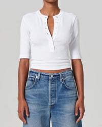 A person wears the Citizens of Humanity Nali Short Sleeve Henley in White, featuring ribbed organic cotton and a partially unbuttoned front, paired with high-waisted blue jeans. The background is plain light gray.