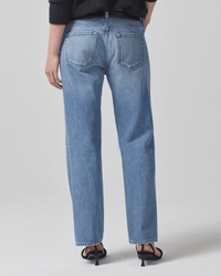 A person faces away, wearing Citizens of Humanity's Neve Low Slung Relaxed in Misty jeans, a black long-sleeve top, and black heeled sandals against a plain light background.