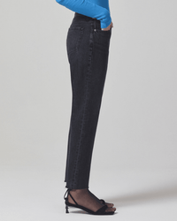 A person stands in profile wearing Citizens of Humanity’s Neve Low Slung Relaxed in Voila black corduroy pants, paired with a bright blue long-sleeve top and black open-toe heeled sandals against a plain light background.
