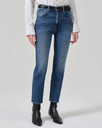 A person stands against a plain light background wearing Citizens of Humanity Zurie Ankle in Abalone jeans, a white striped button-up shirt, black belt, and black ankle boots. Head is out of frame.