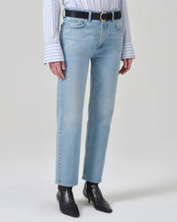 A person wears Citizens of Humanity straight leg jeans, a striped button-down shirt with long cuffs, a black belt with a gold buckle, and Zurie Ankle in Garland boots by Citizens of Humanity, standing against a plain light background.