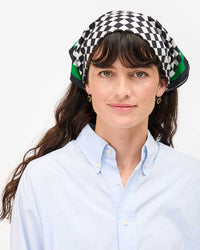 Woman with long brown hair wears a Clare V. Cream & Black Checker Bandana—a versatile accessory—paired with a light blue button-up shirt, looking at the camera and smiling slightly against a plain white background.