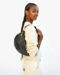 A woman with long braids, in a cream sweater and light pants, smiles while holding the Clare V. Grande Fanny in Black Nouveau with an adjustable strap against a plain white background.