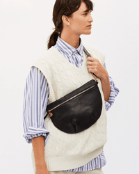 A person wears a cream cable-knit sweater vest over a blue and white striped shirt, carrying the Clare V. Grande Fanny in Black Perf crossbody bag, standing against a plain background.
