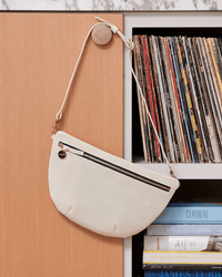 The Clare V. Grande Fanny in Cream Perf, a cream-colored textured perforated leather bag with a zipper, hangs on a wooden knob beside a shelf of vinyl records and books.