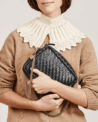 A woman in a tan knit sweater with a white collar holds the Clare V. Midi Sac in Black Woven Zig-Zag, featuring a gold chain strap, against her chest.
