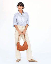 A person stands against a white background wearing a light blue and white striped shirt, cream-colored pants, tan flats, and holding the Clare V. Moyen Messenger in Natural Woven Checker.