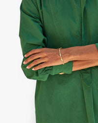 A person in a green long-sleeve shirt stands with arms crossed, showcasing the Clare V. Sardine Link Bracelet in Vintage Gold on their wrist against a plain white background.