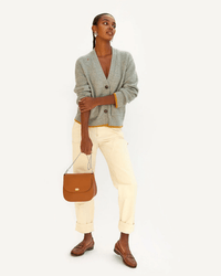 A woman stands against a plain background in a grey cardigan, cream pants, and brown loafers, holding a Clare V. bag with the Snake Chain Shoulder Strap in Silver and looking slightly upward with a relaxed expression.