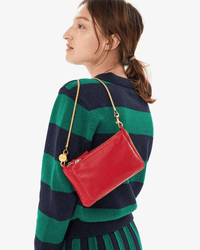 A person with brown hair in a ponytail wears a navy and green striped sweater and matching skirt, carrying a Clare V. bag with the Snake Chain Shoulder Strap in Vintage Gold over their shoulder.