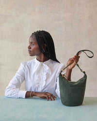 A woman in a white blouse with a bow collar sits at a table, looking to the side and holding up the Clare V. Suede Petite Jeanne in Loden, which features a customizable strap.
