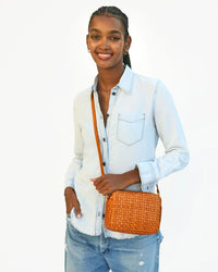 A woman in a light blue denim shirt and ripped jeans smiles while holding the Clare V. Woven Checker Midi Sac in Natural, with one hand in her pocket, standing against a plain white background.
