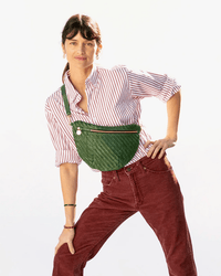 Wearing a red and white striped shirt, red pants, and the Clare V. Woven Zig-Zag Grande Fanny in Evergreen with an adjustable strap, a person poses with one hand on their hip against a plain background.