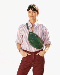 A woman in a red-and-white striped shirt carries the Clare V. Woven Zig-Zag Grande Fanny in Evergreen, featuring a green woven leather design and adjustable strap, while smiling at the camera against a plain background.