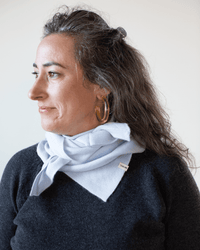 A woman with long brown hair, large gold hoop earrings, and a black top wears the COLORI Large Triangle Bandana in Porcelain as she looks to the left against a plain background.