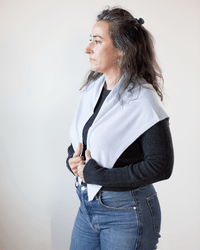A woman with long, wavy hair, in a black long-sleeve top and blue jeans, stands against a light background looking to the side, wearing the COLORI Large Triangle Bandana in Porcelain draped as a shawl over her shoulders.