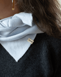 Close-up of a person in a light blue COLORI hoodie with a visible “COLORI” tag, layered under a dark sweater. The person’s hair, hoop earring, and the COLORI Large Triangle Bandana in Porcelain are also partially visible.