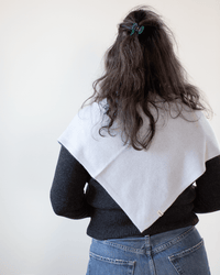 A person with long dark hair, partially tied up, wearing a dark sweater, blue jeans, and a light gray shawl draped over their shoulders, completes the look with COLORI’s Large Triangle Bandana in Porcelain, seen from behind against a plain background.