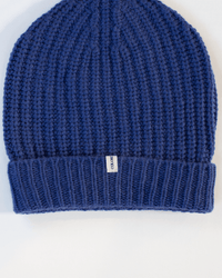 A close-up of the Ribbed Beanie in Neptune by COLORI shows its blue ribbed knit, folded brim, and a small white tag with the brand name. The cozy winter hat is set against a plain white background.