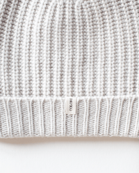 Close-up of the Ribbed Beanie in Stucco by COLORI, featuring a soft grey knit texture and a small white COLORI tag sewn at the edge. The image showcases the ribbed pattern and cozy winter style.