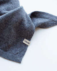 A close-up of the Small Triangle Bandana in Kensington by COLORI, featuring soft gray 100% cashmere knit fabric and a small beige COLORI tag sewn into the seam, set against a white background.