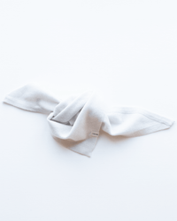 The COLORI Small Triangle Bandana in Stucco, a soft light gray fabric, is laid flat and loosely knotted at the center on a white background, with a small white tag visible near the knot.