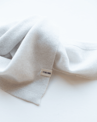 The Small Triangle Bandana in Stucco by COLORI, a soft and light gray accessory, rests on a white surface. A small white tag displaying the brand name COLORI in black text is sewn onto the fabric.