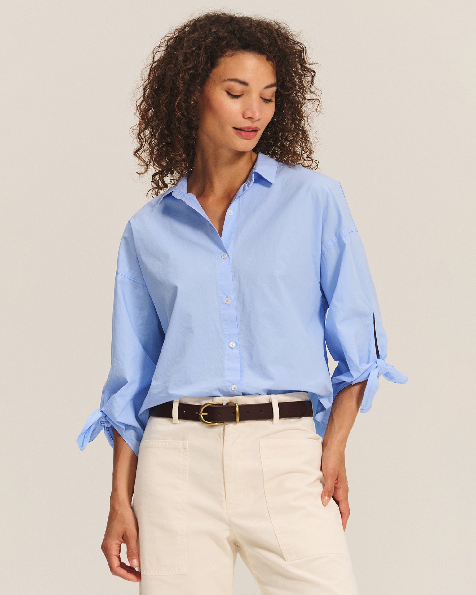 A woman with curly hair wears the Velvet by Graham & Spencer Arlette Knot Sleeve Button Up in Chill and off-white pants, accessorized with a black belt. She looks down slightly, standing against a plain light background.