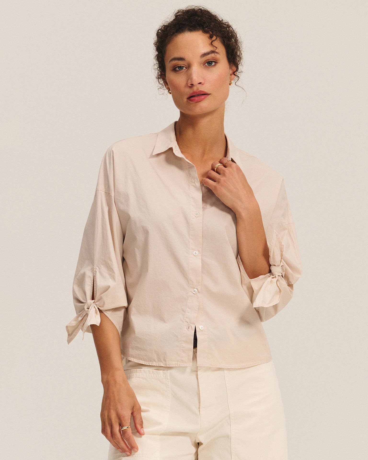 A woman with curly hair wears the Arlette Knot Sleeve Button Up in Sesame by Velvet by Graham & Spencer, paired with white pants. She stands against a plain background, gazing at the camera with a neutral expression.