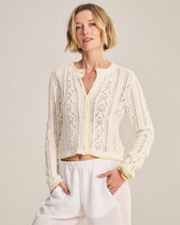 A person with short blonde hair wears the Velvet by Graham & Spencer Daisy Long Sleeve Crew Cardi in Ivory with loose white pants, standing confidently with one hand in their pocket against a neutral background.